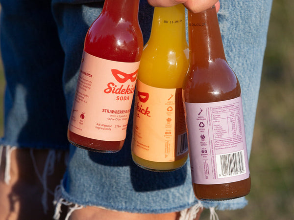 Hand holding three Sidekick glass bottles — Strawberry & Plum, Mango, and nutrition label view
